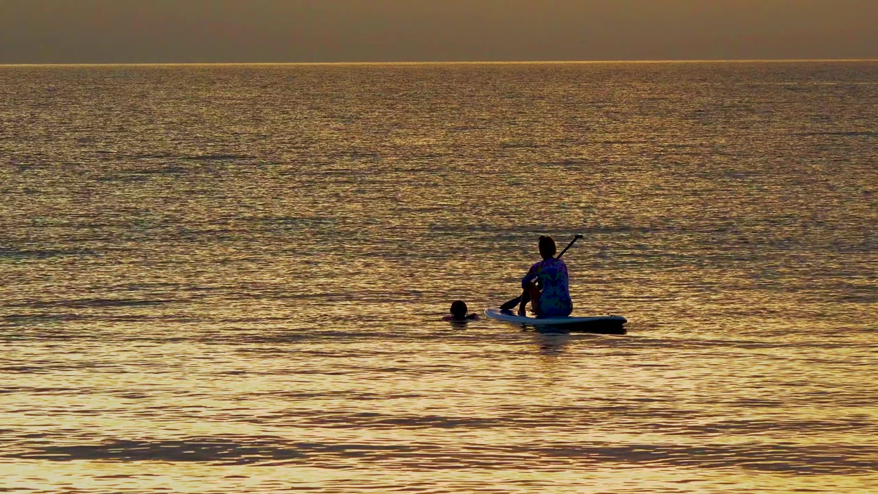 The kayaker appears farther to the right with the swimmer behind on golden ocean water in El Sargento La Ventana, Mexico, under a steady horizontal skyline.