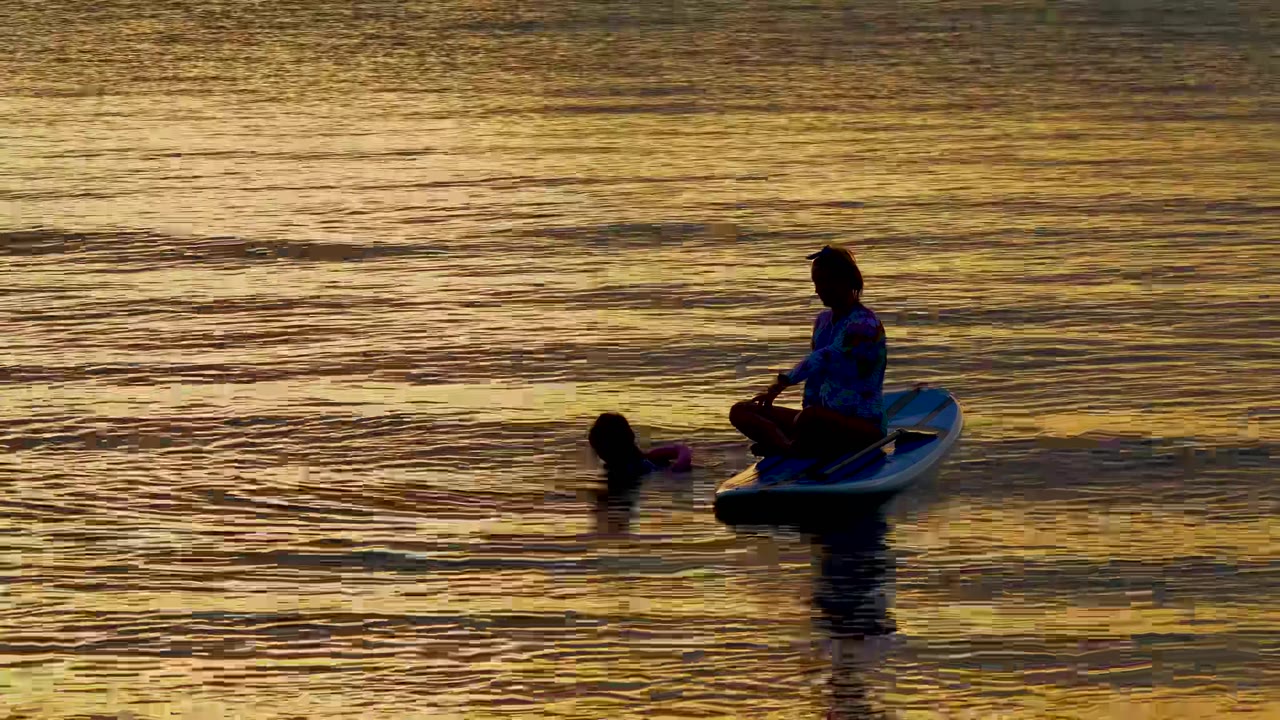In El Sargento, Mexico, a silhouetted person sits on a paddleboard while another person treads water nearby in rippled golden light.