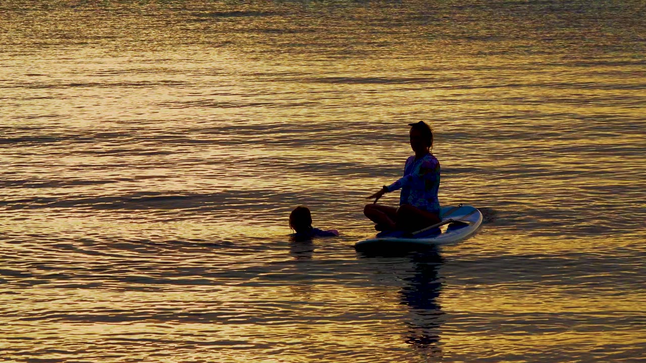 Silhouetted paddleboarder and swimmer on calm reflective water.