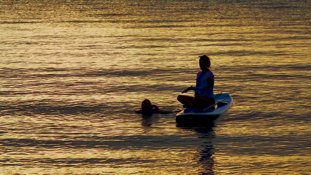 A dark swimmer floats left of a seated paddleboarder, with broad bands of gold and darker ripples filling the El Sargento water behind them.