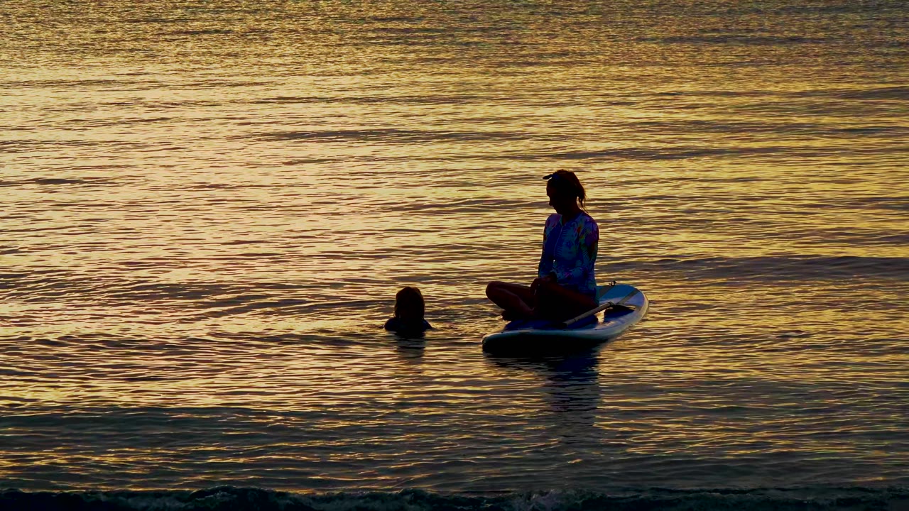 At El Sargento, Mexico, a silhouetted person sits cross-legged on a board while a swimmer's head appears nearby in gold sunset water.