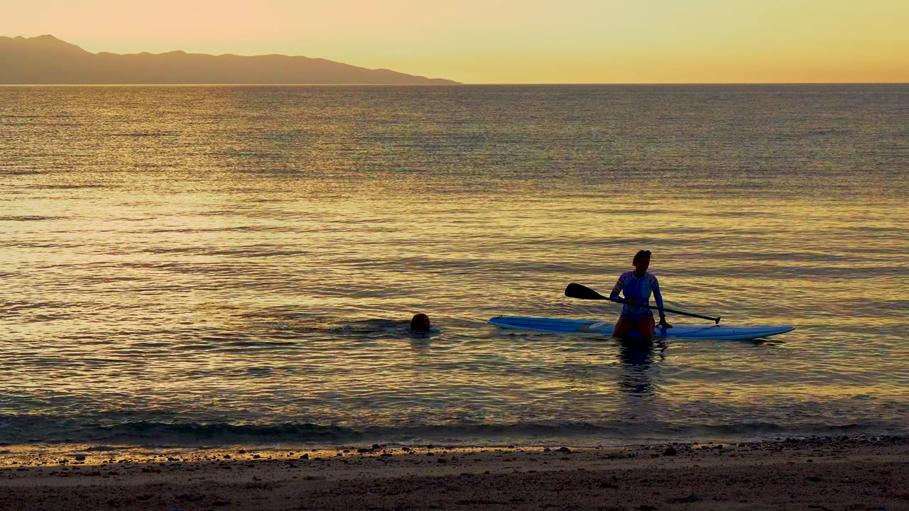 Two people are in the ocean near the sandy beach in El Sargento, Mexico, with one swimmer near the board and golden sunset light across the water.