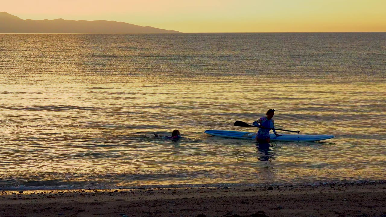 A person holds a blue paddleboard in shallow ocean water in El Sargento, Mexico, while another person swims nearby under warm sunset reflections.