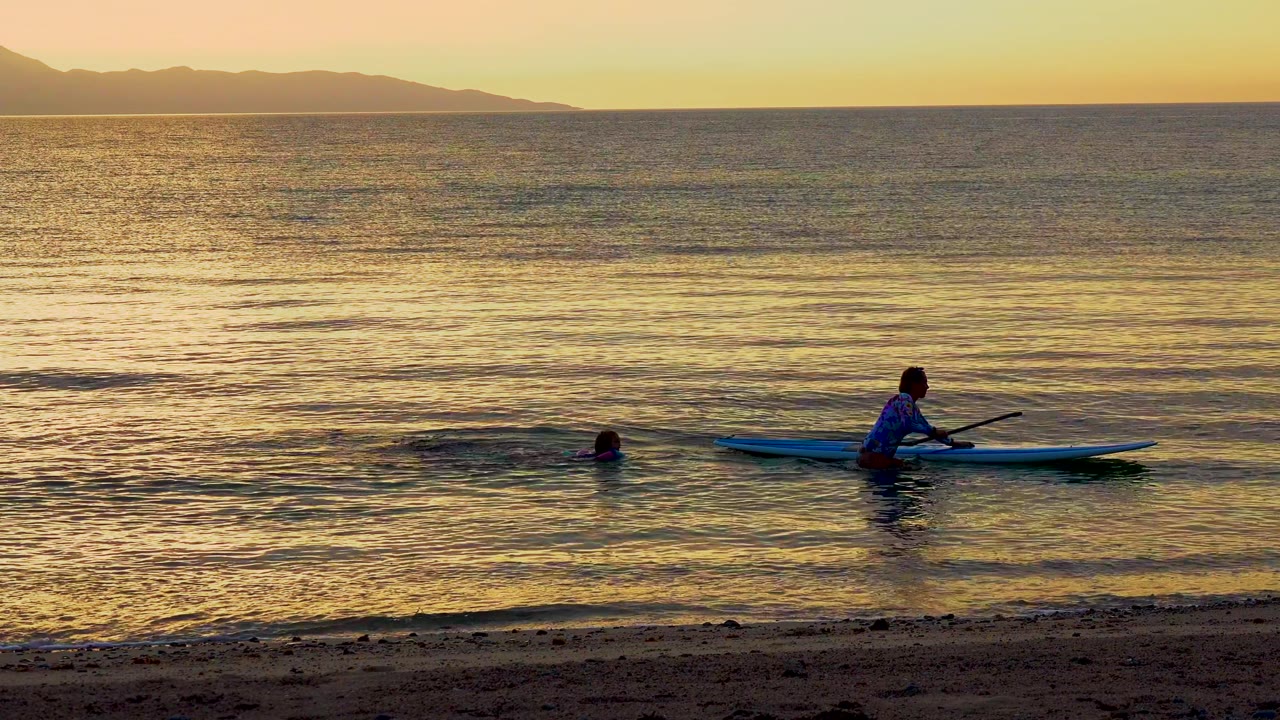 In El Sargento, Mexico, the paddleboarder is farther to the right of frame while the swimmer remains in the water, with low waves and a mountain silhouette beyond the ocean.