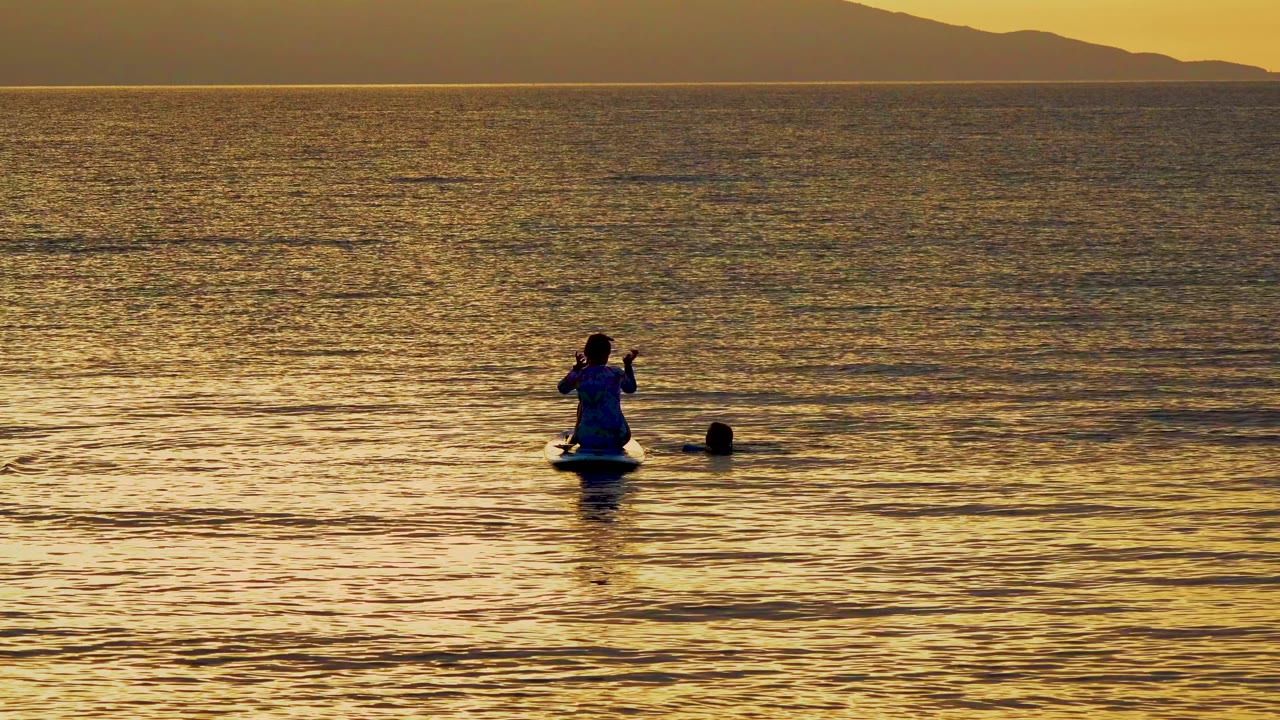 A silhouetted paddleboarder with a paddle and a nearby swimmer sit low in golden sunset water off El Sargento, Mexico, with mountains on the horizon.