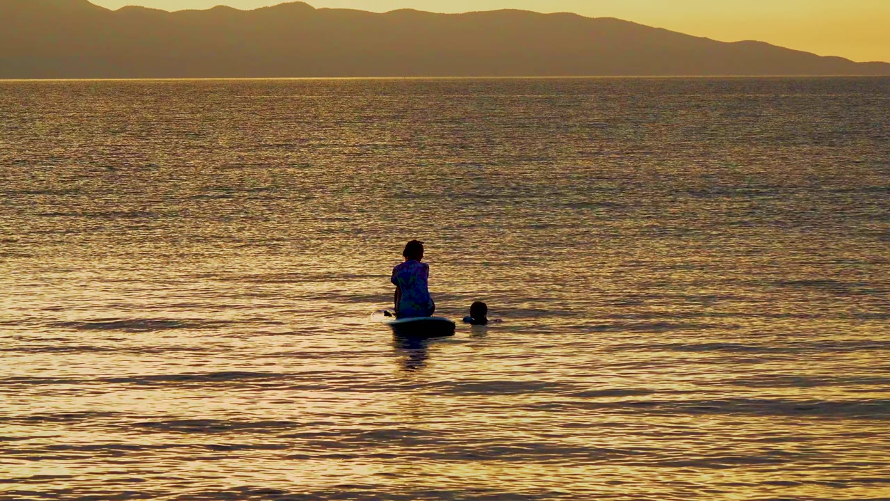 Seated paddleboarder and nearby swimmer shape in calm golden water.