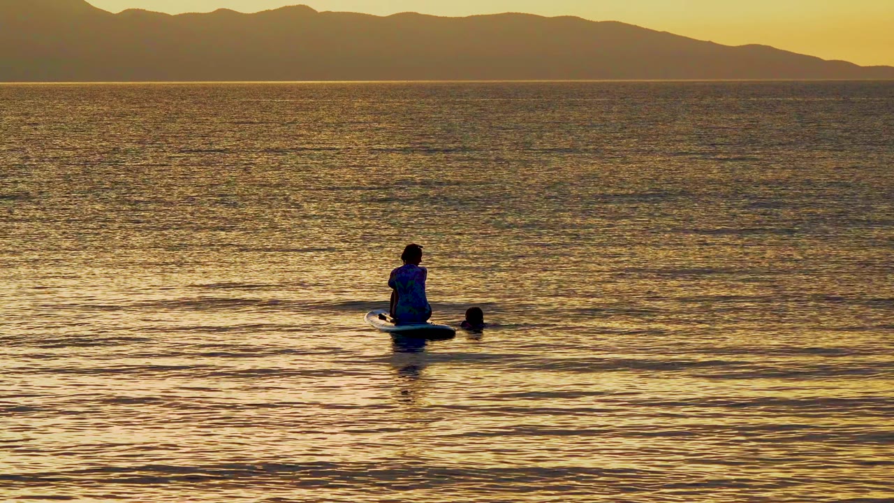 At 0:02 in El Sargento, Mexico, the paddleboarder sits farther right in the wide frame, with the small nearby figure separated by a strip of darker ripples.
