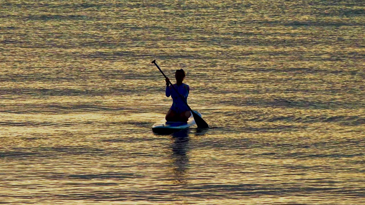 A silhouetted paddler kneels on a paddleboard in El Sargento, Mexico, with the paddle lowered beside the board on calm golden water.