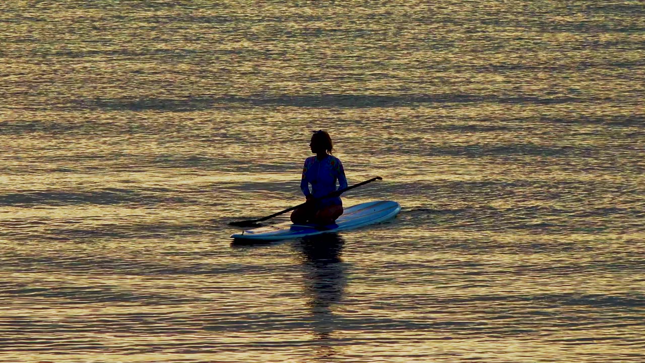 A person sits on a blue paddleboard in El Sargento, Mexico, with a paddle held across the board as golden water fills the frame.