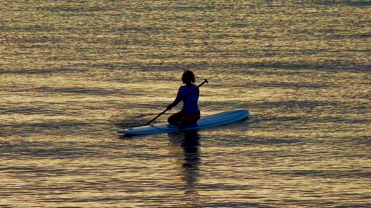 A silhouetted person on a blue paddleboard in El Sargento, Mexico, plants the paddle beside the board amid broad bands of golden reflected light.