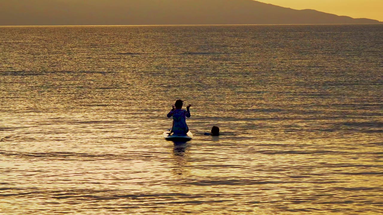 A paddleboarder sits in calm golden water near a swimmer in El Sargento, Mexico, with a low mountain silhouette on the horizon.