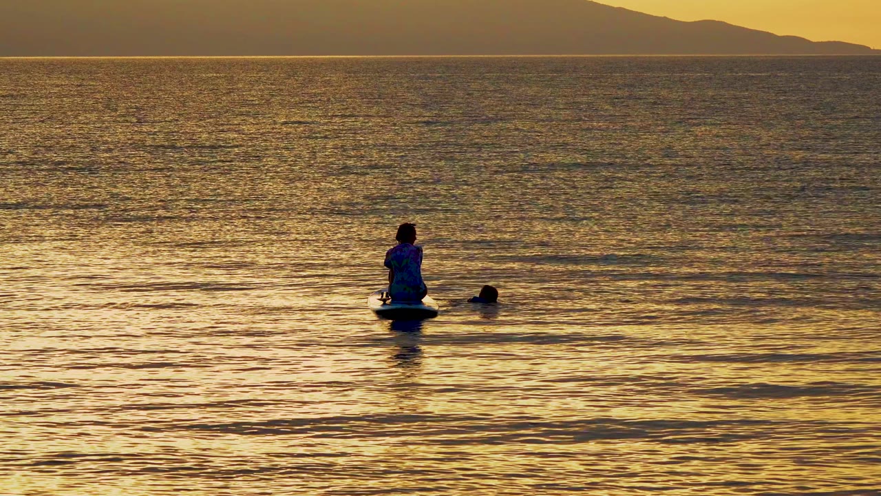 A seated paddleboarder and nearby swimmer sit slightly farther right in the frame, backed by golden water and faint mountains in El Sargento, Mexico.