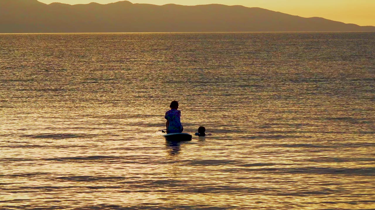 Backlit person and dog float on a paddleboard in calm ocean water.