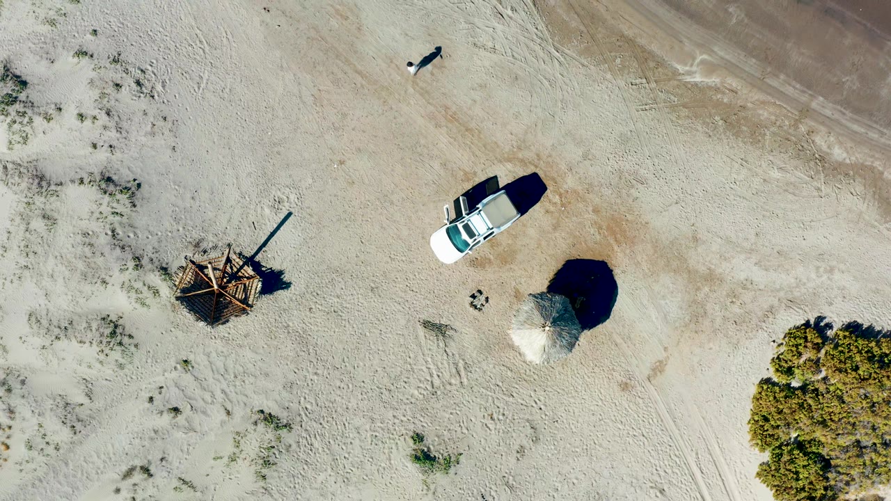 Overhead frame from Juncalito, Baja California Sur showing a white car on pale sand near scrub, a rough wooden structure, a round shade structure, and a small distant person near the top edge.