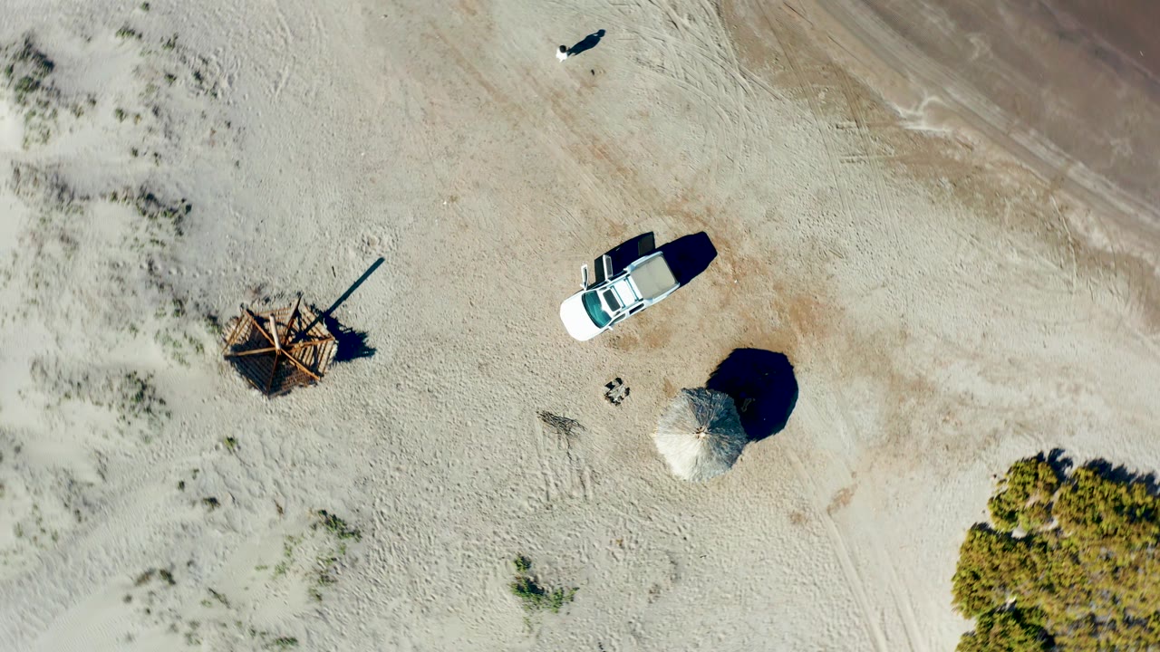 Near-vertical aerial view in Juncalito with a white vehicle beside pale sand, low scrub, a round shade structure, a rough wooden structure, and a small distant person along the upper sandy area.