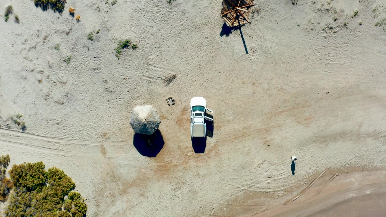 Overhead frame in Juncalito, Baja California Sur showing a white car with an open door beside a thatched umbrella, with a small person walking across pale sand near shrubs.