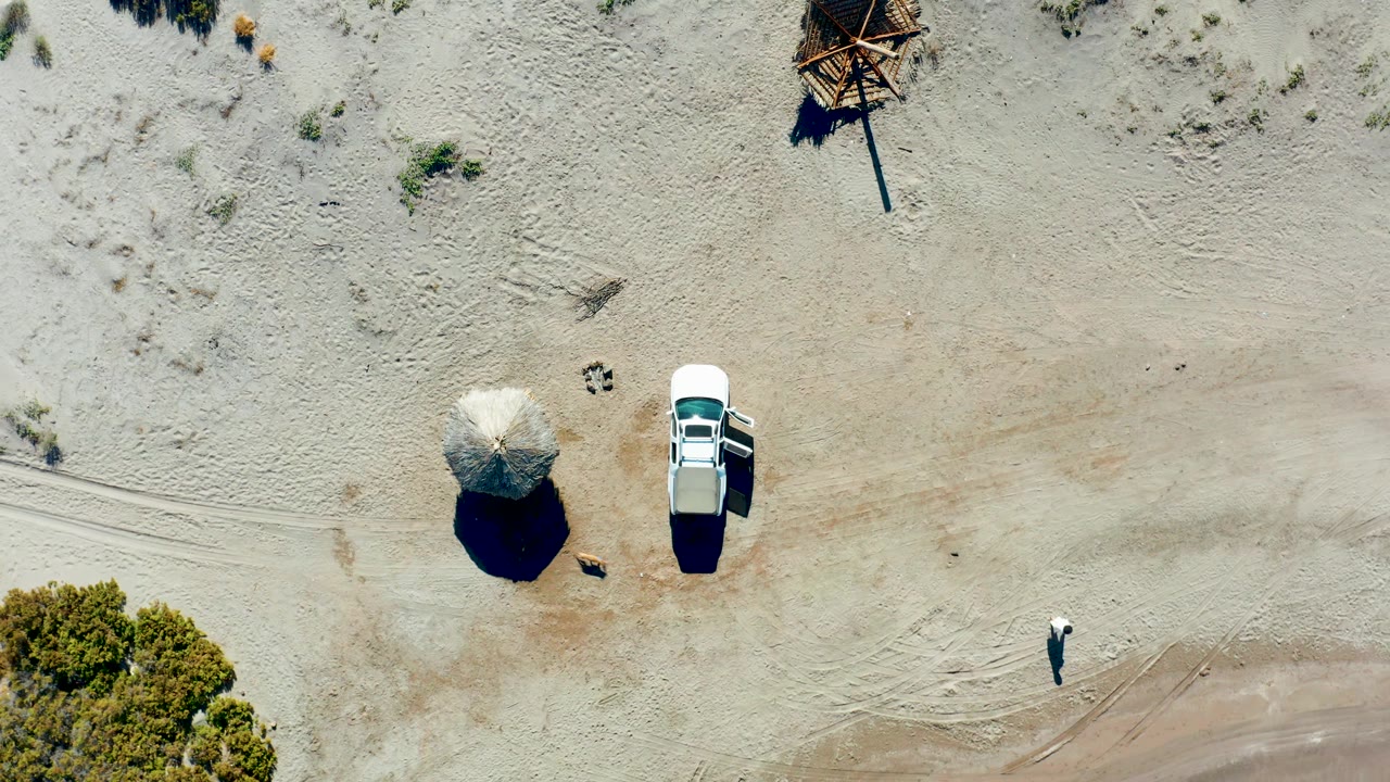 White car with open door beside a thatched umbrella as a person walks across sand.