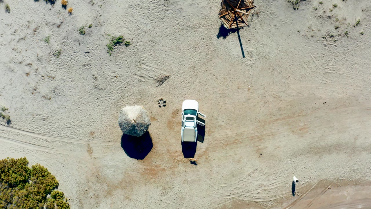 Top-down frame in Juncalito, Baja California Sur showing the white car and umbrella centered against pale sand, with the person now closer to the lower right edge.