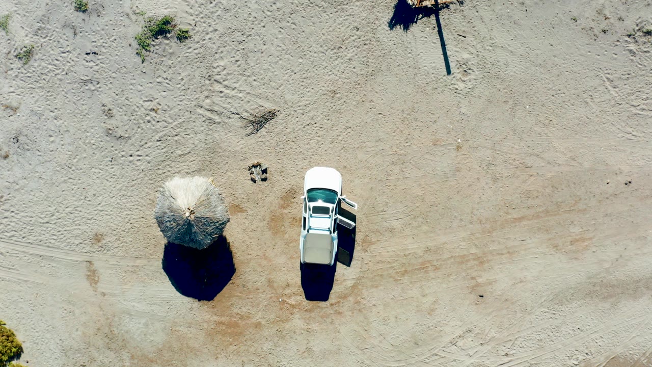 Overhead view in Juncalito, Baja California Sur, with a white car near a round thatched beach umbrella on pale sand and no visible people.