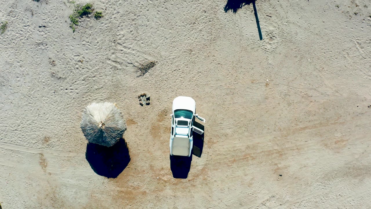 Aerial frame in Juncalito, Baja California Sur, showing the white car beside the thatched umbrella as strong sun defines their dark shadows on sand, with no visible people.