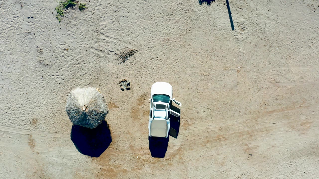 Top-down Juncalito beach frame with the white car's rear area visible, the thatched umbrella to its left, faint tire marks in the sand, and no visible people.