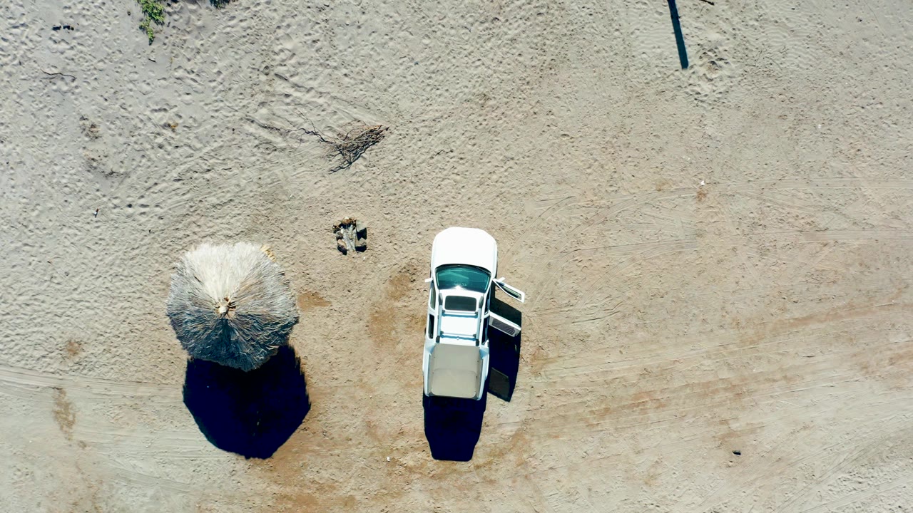 Overhead view in Juncalito, Baja California Sur, with the thatched umbrella casting a round shadow near the white car as the trunk action continues and no people are visible.