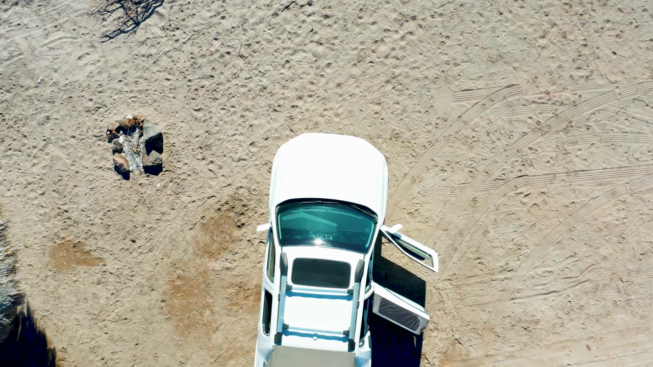 At 0:02 in Juncalito, the aerial frame shows the white car at the lower edge, its open door casting a dark shadow, and the person separated above on the sand.