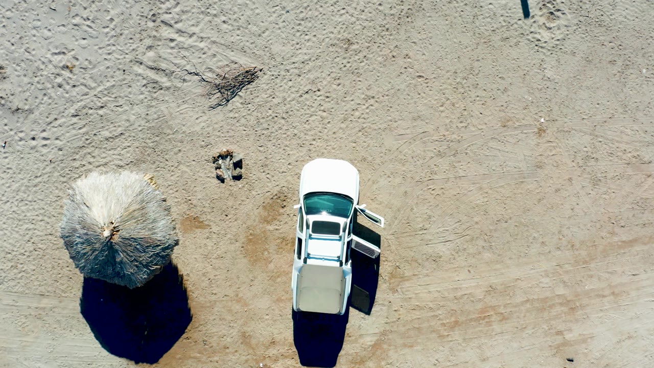 Top-down aerial frame in Juncalito, Baja California Sur, Mexico, showing a white car with an open side door beside a thatched beach umbrella on pale sand.