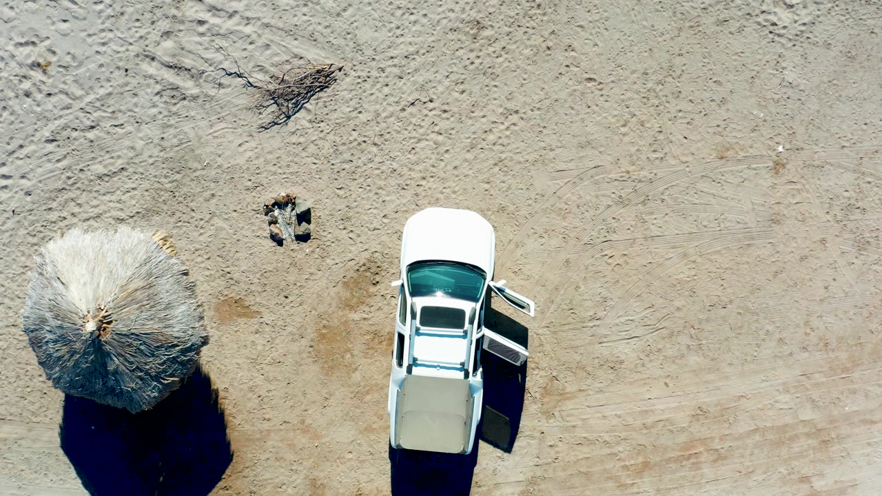 Top-down frame from Juncalito, Baja California Sur, Mexico, with the white car near the right edge, an open door, a thatched umbrella to the left, and textured sand around them.