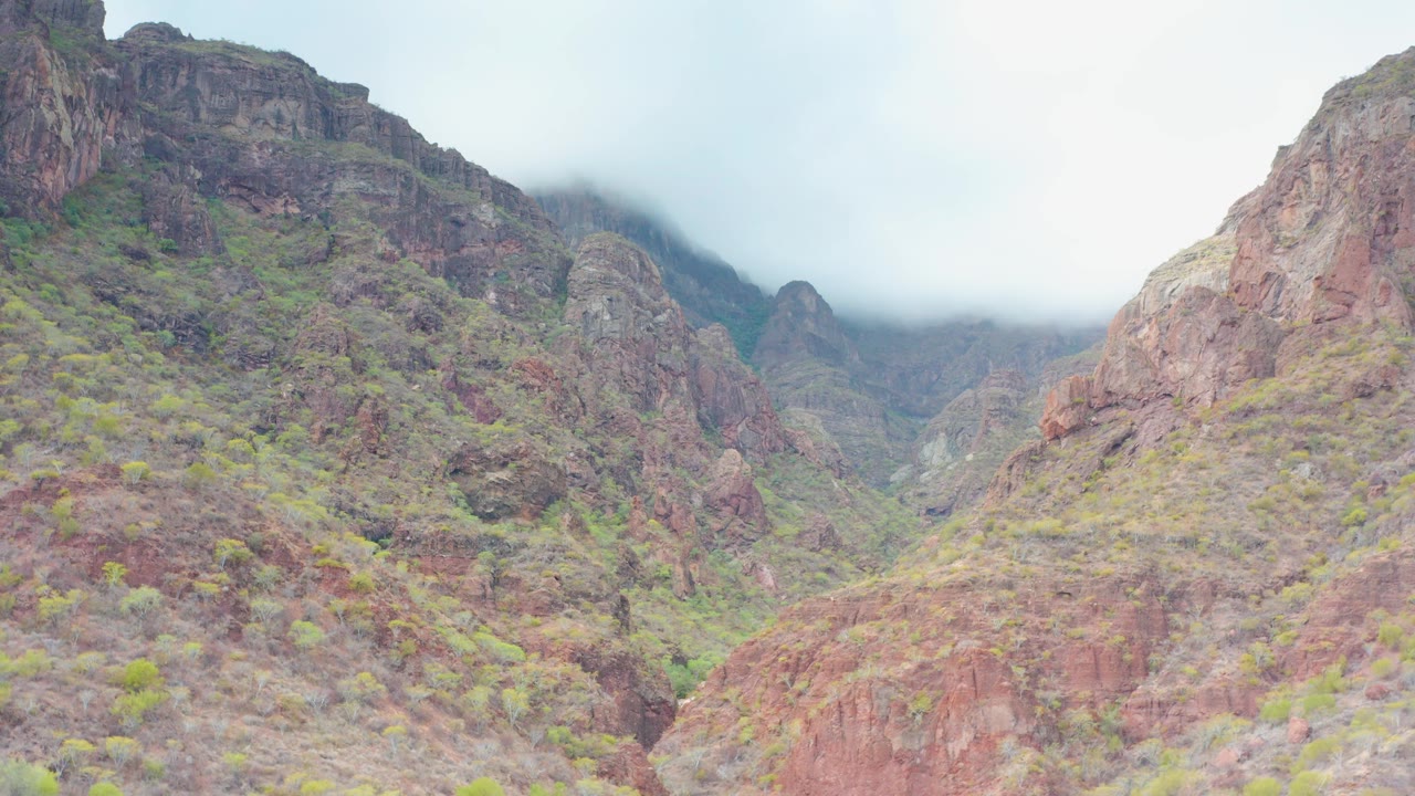 Opening frame of a Puerto Escondido, Baja California Sur red rock mountain valley with low mist on the upper ridges, sparse green vegetation, and no visible people.