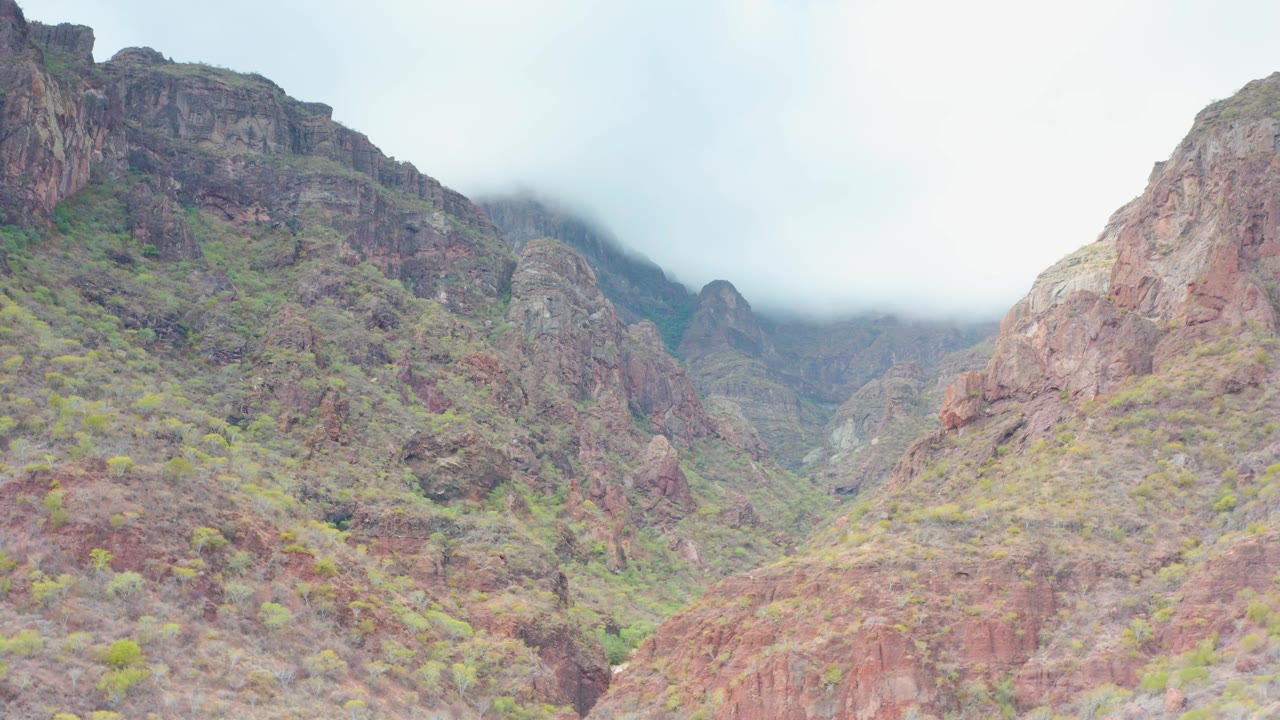 Early frame showing the central Puerto Escondido, Baja California Sur valley framed by reddish cliffs, green shrubs, low cloud, and no visible people.