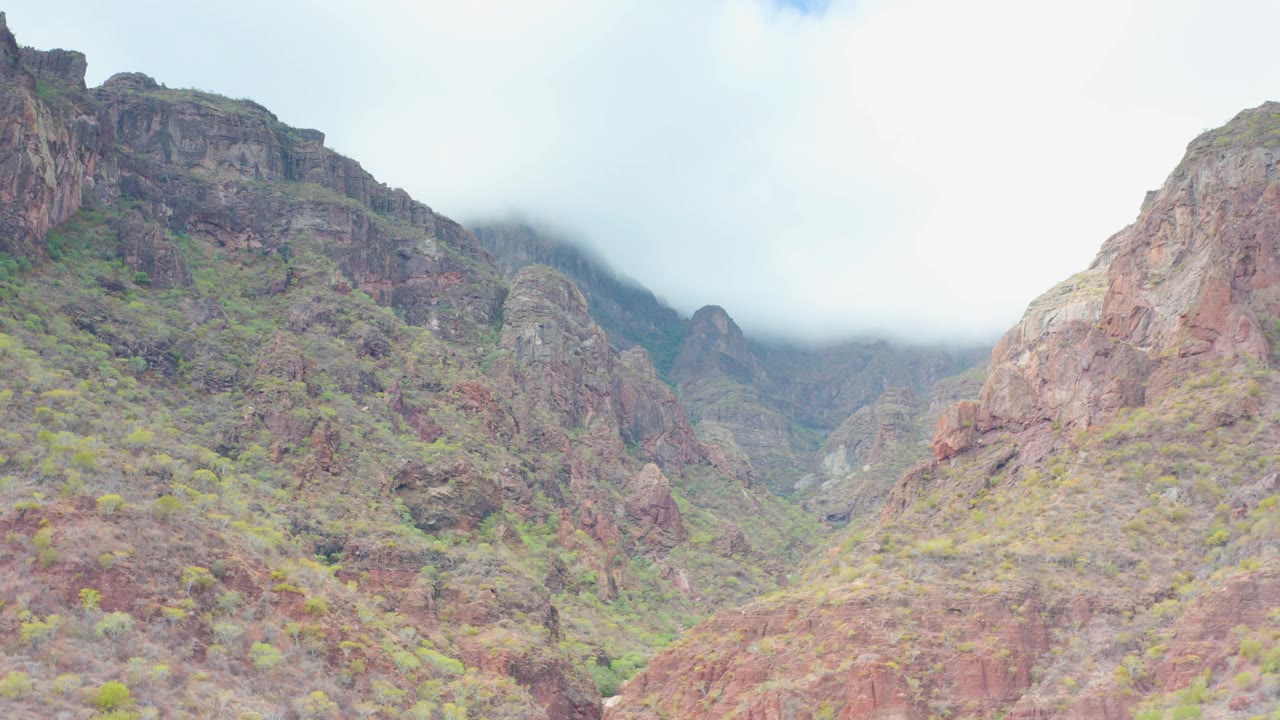 Mid-early frame of rugged red and brown mountain walls near Puerto Escondido, Baja California Sur, with pale sky, misted upper slopes, scattered greenery, and no visible people.