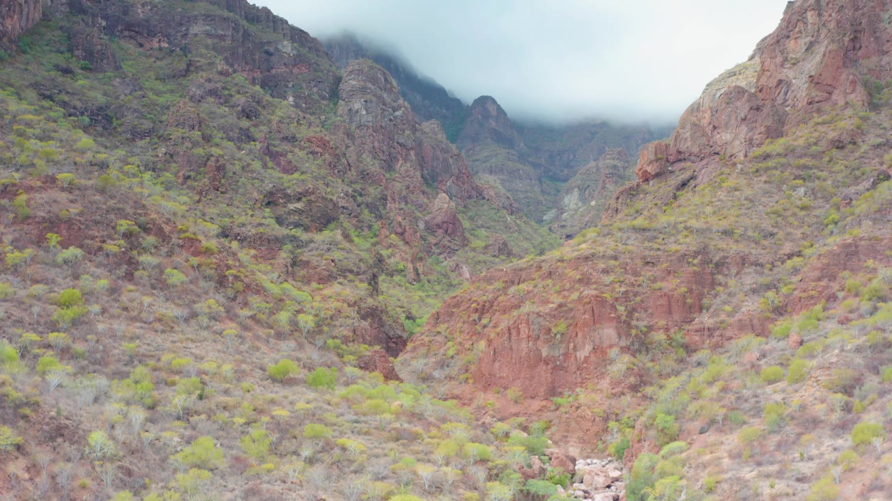 A steep red rock wall in Puerto Escondido, Baja California Sur rises behind scrub-covered canyon terrain, with low mist hiding parts of the upper ridgeline and no visible people.