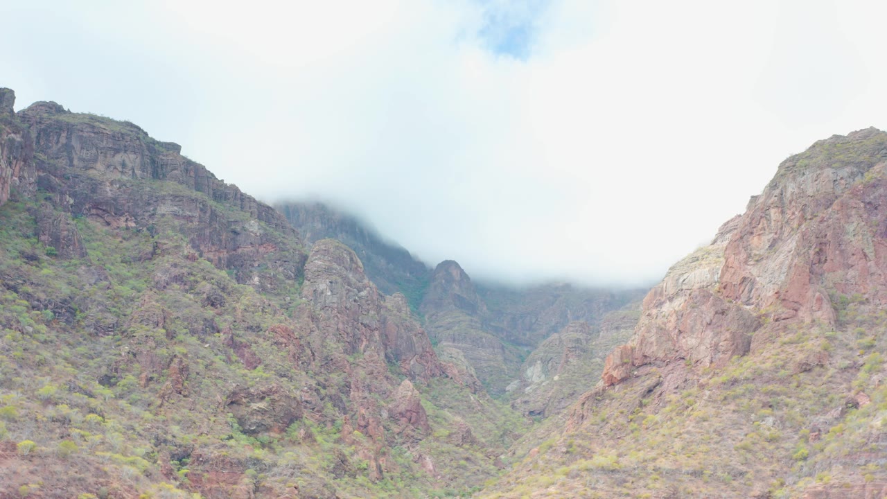 A central red-brown rocky slope rises below pale cloud in Puerto Escondido, Baja California Sur, with sparse vegetation and no visible people.