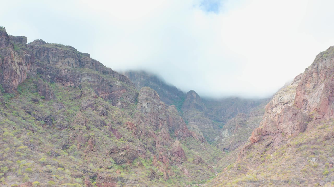 Wide view of a rocky mountain valley in Puerto Escondido, Baja California Sur, with green vegetation on red-brown slopes, low mist along the upper ridge, and no visible people.