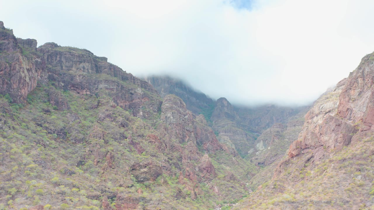 A central rocky peak rises above scrub-covered slopes in Puerto Escondido, Baja California Sur, while pale cloud and mist obscure parts of the higher terrain, with no visible people.