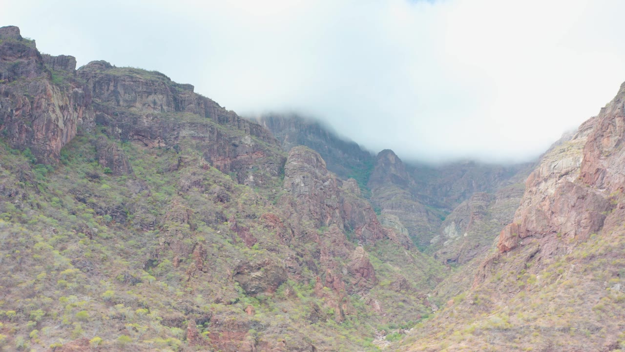 The frame holds on steep cliffs and layered mountain slopes in Puerto Escondido, Baja California Sur, with low cloud sitting on the ridge and no visible people.