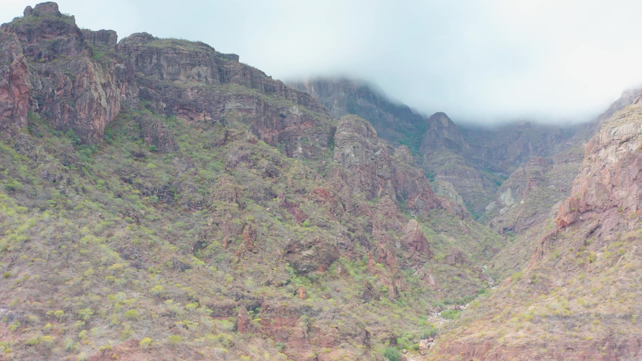 Opening view of rocky cliffs and green-brown slopes in Puerto Escondido, Baja California Sur, with cloud hanging over the upper ridges and no visible people.
