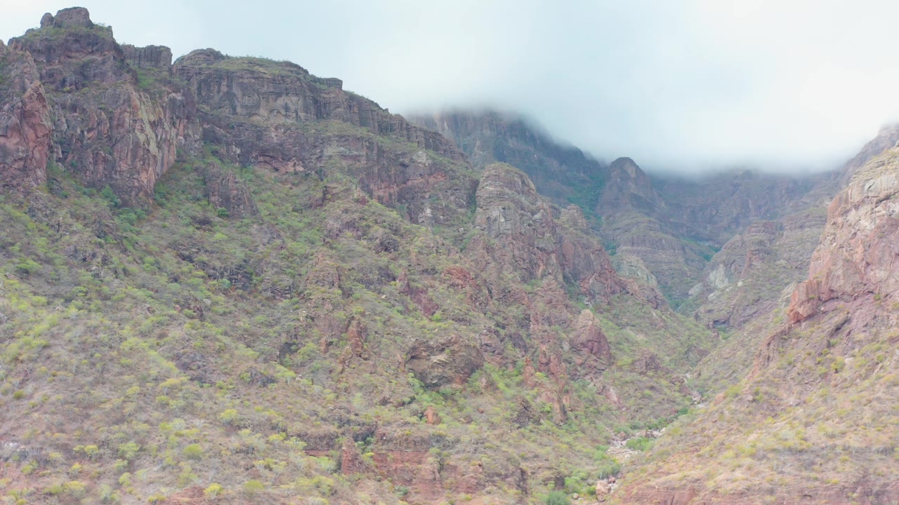 A rugged cliff face and layered mountain valley in Puerto Escondido, Baja California Sur, sit beneath pale low cloud, with no visible people or structures.