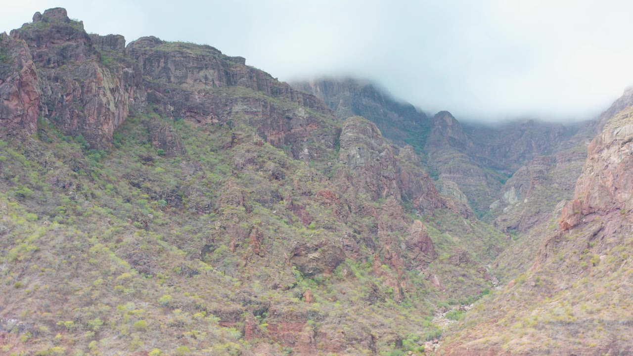 The frame shows steep rocky slopes with patches of green vegetation in Puerto Escondido, Baja California Sur, while mist obscures the higher peaks and no people are visible.