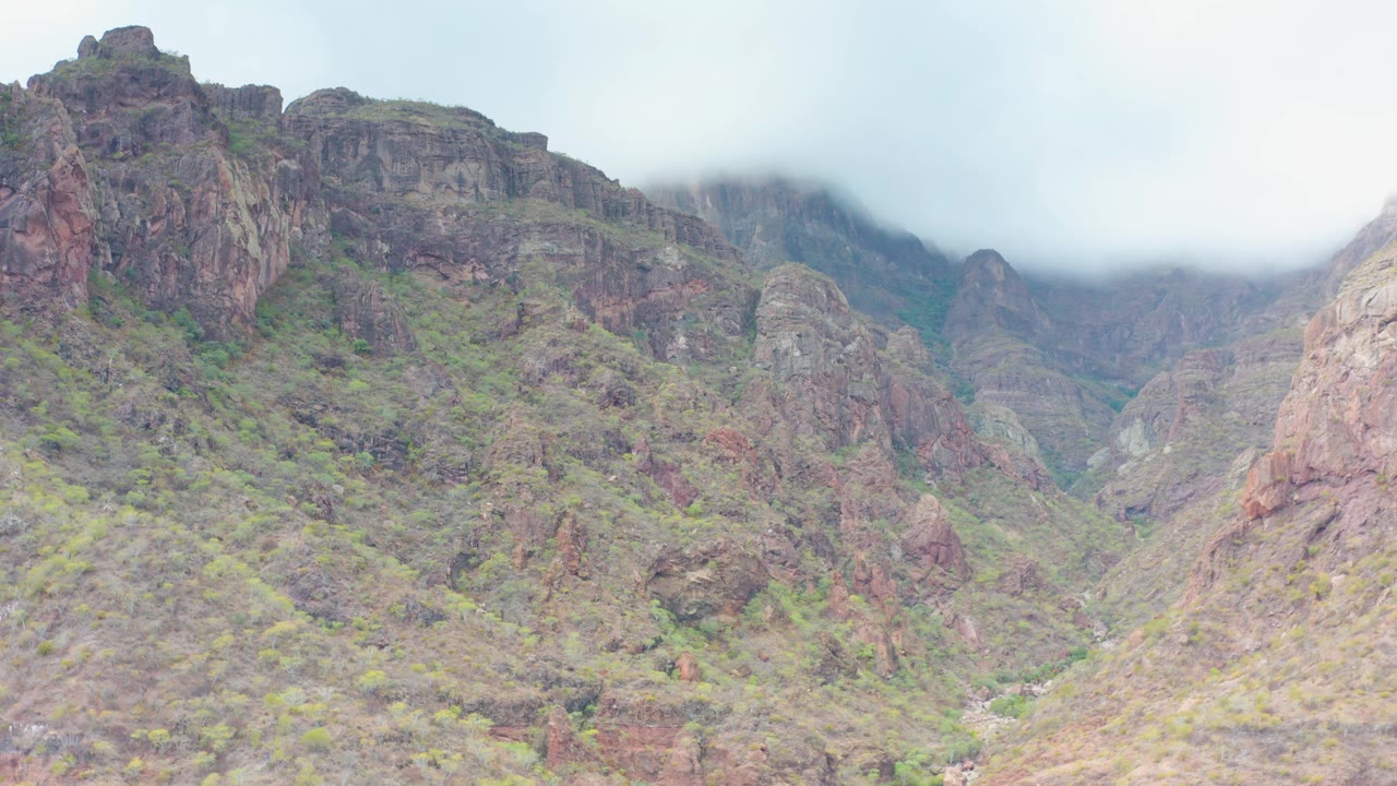 A slightly lower view of the rocky Puerto Escondido mountain valley shows jagged formations, pale overcast sky, and low cloud across the upper background, with no visible people.