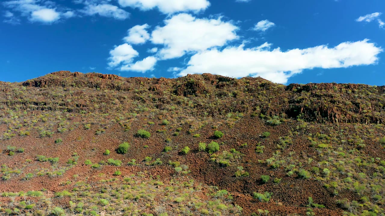 Third frame holds on a rough San Javier ridge with dark rocky outcrops, patchy green vegetation, a saturated blue sky, and no visible people.