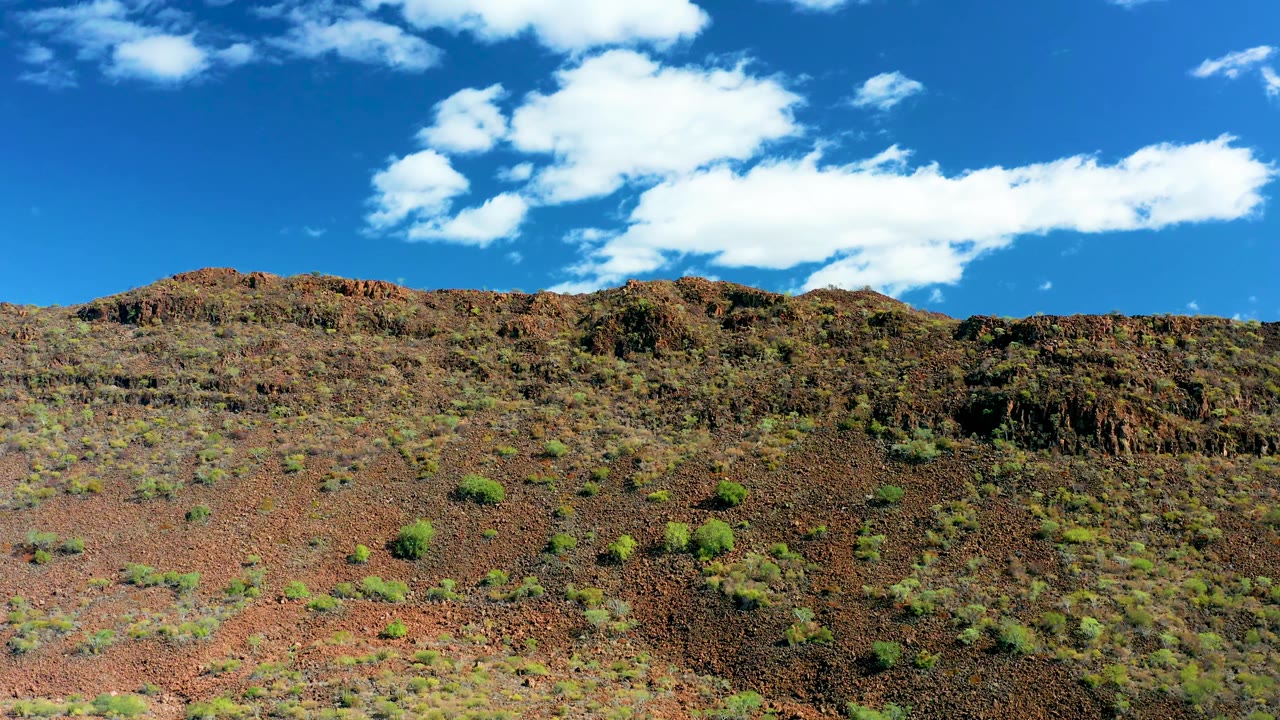 Rocky red-brown hillside with sparse shrubs below blue sky and white clouds.