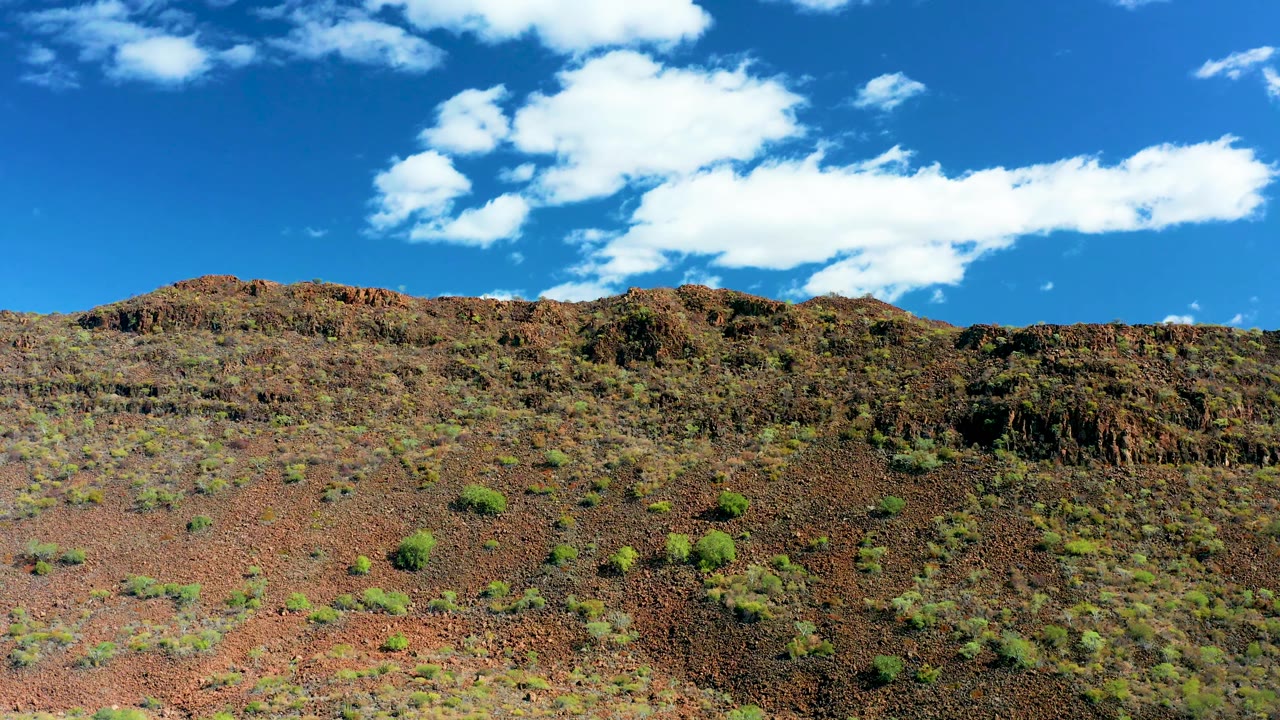 San Javier hillside frame with red-brown rock, sparse green shrubs, a rugged ridge line, a large white cloud across the blue sky, and no visible people.