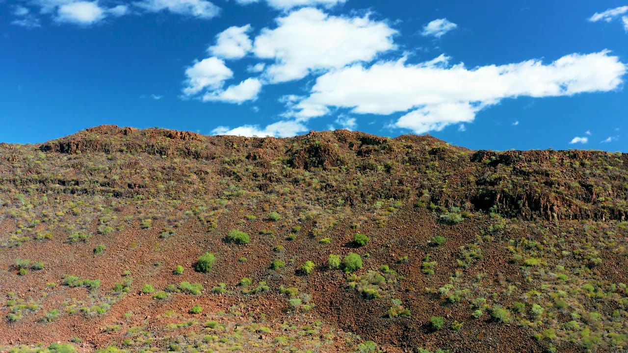 San Javier, Baja California Sur landscape frame with red rocky ground, low sparse shrubs, a dark rugged ridge, white clouds, and no visible people.