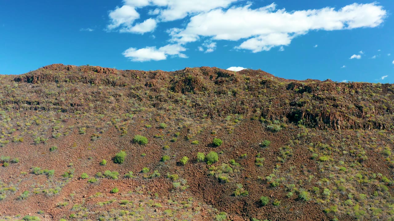 At 0:03, a rocky desert hillside in San Javier fills the lower frame, with scattered green shrubs, a clean blue sky, white clouds, and no visible people.