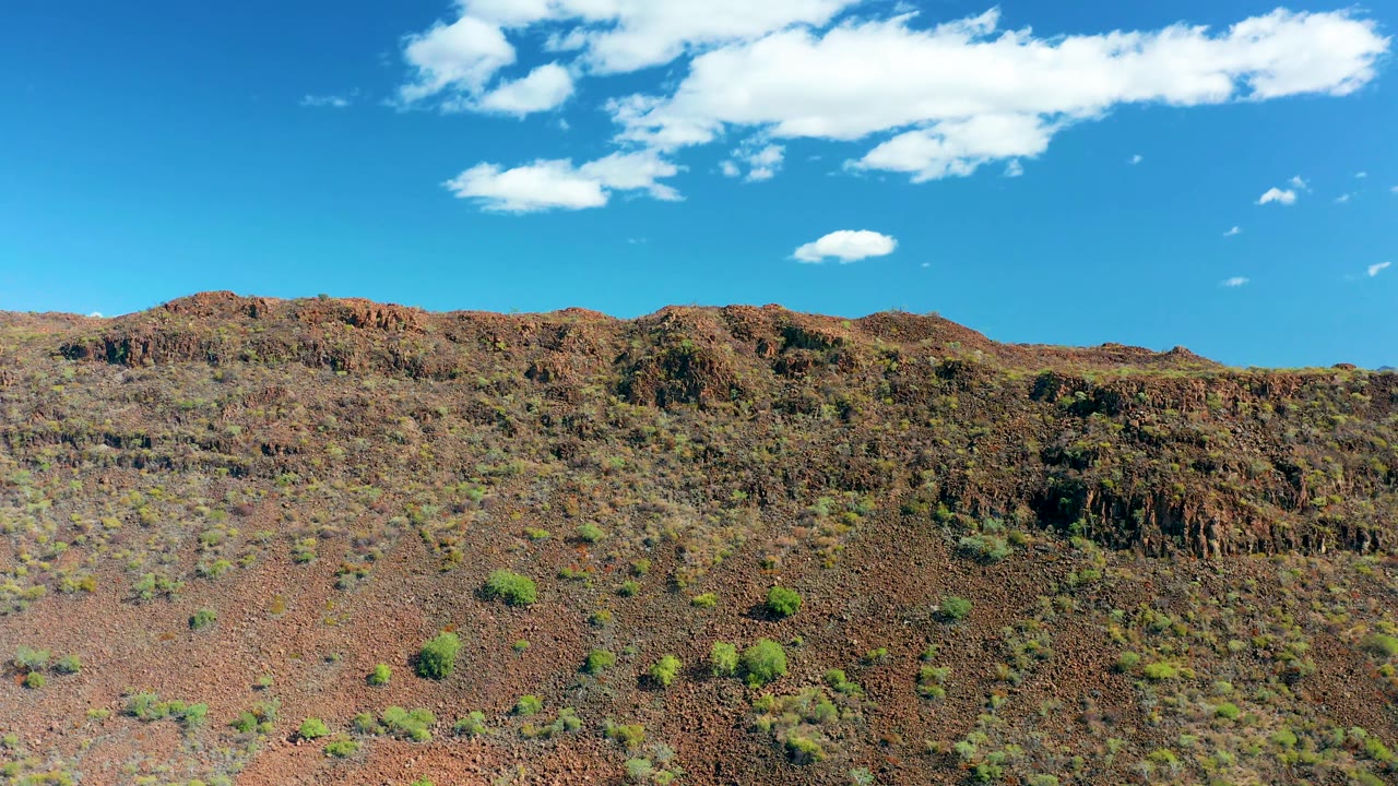 Opening frame of San Javier, Baja California Sur, showing a reddish rocky hillside with sparse green shrubs below a bright blue sky, with no visible people.
