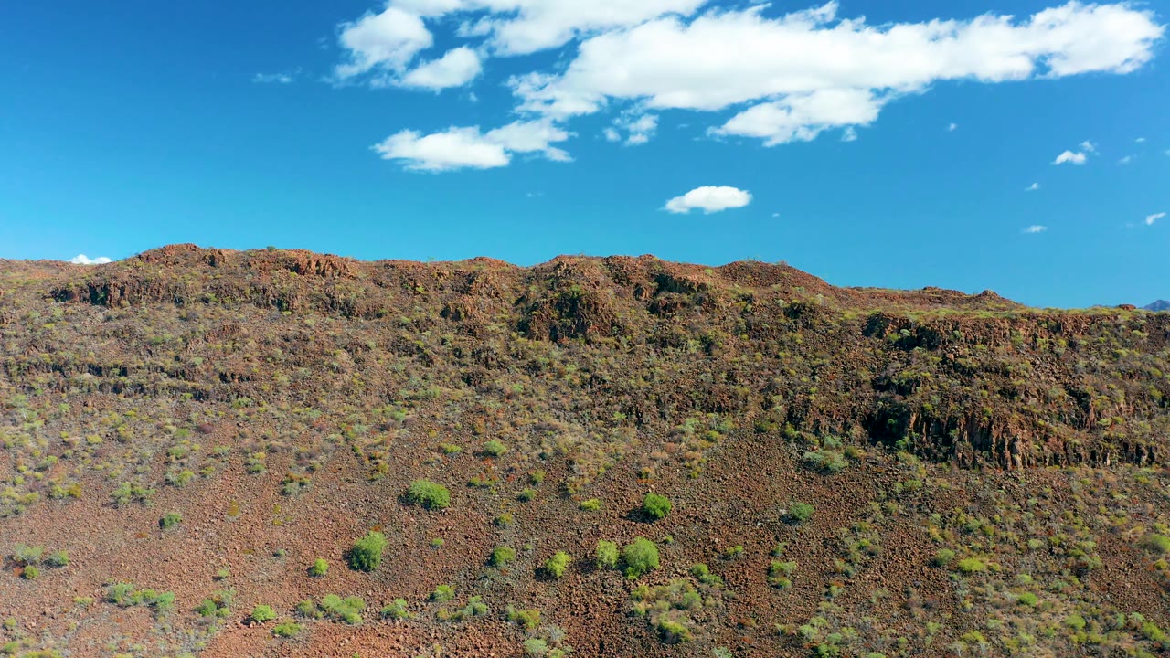 San Javier desert slope continues across the frame, with low green shrubs on reddish ground and small clouds above the ridge, with no visible people.