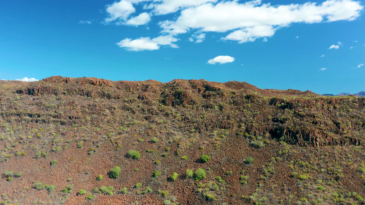 A wider San Javier hillside view shows reddish-brown rock, scattered shrubs, bright cloud patches, and distant mountains beginning to show at the right edge, with no visible people.