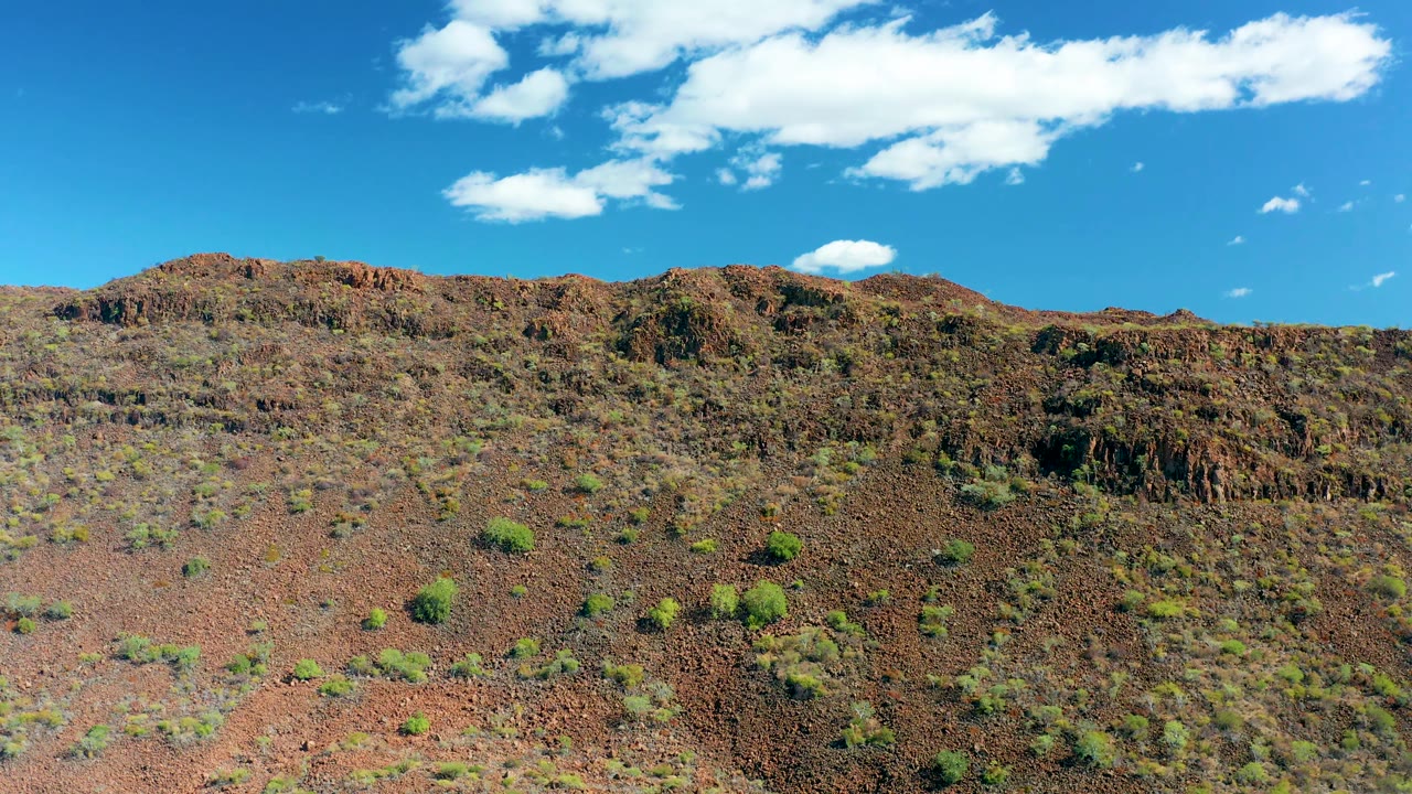 Opening frame of a reddish rocky hillside in San Javier, Baja California Sur, with scattered green shrubs, a low ridgeline, blue sky, and no visible people.